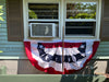 A patriotic bunting banner with red, white, and navy blue stars and stripes hangs in front of a window on a house exterior.